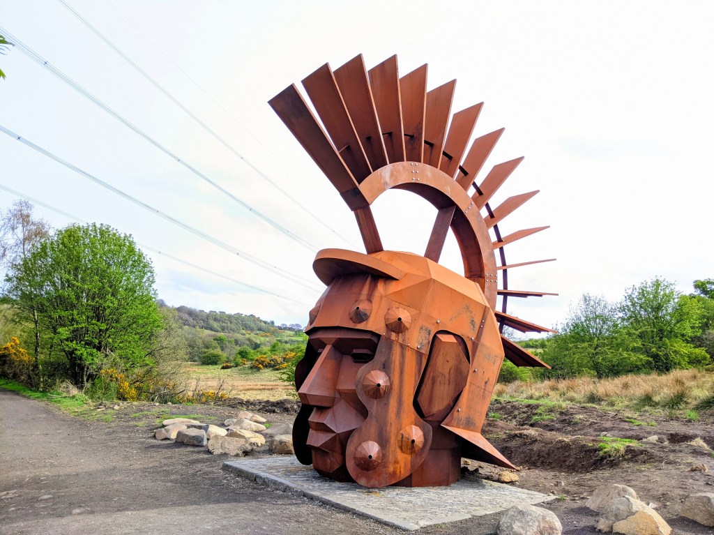 Iron sculpture of head of a Roman soldier sitting on a concrete base beside a path.